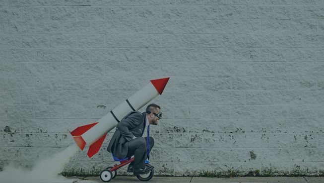 grown man riding tricycle with large rocket on his back 